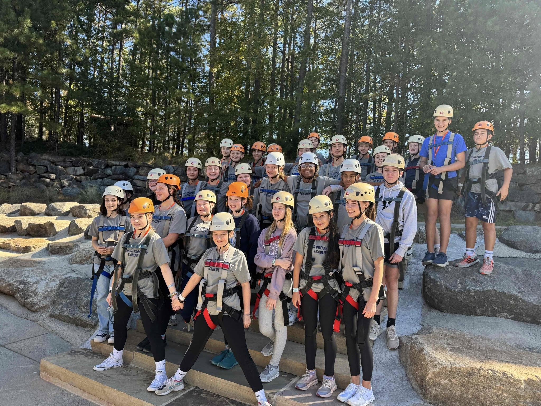 Group of children and adults wearing helmets outdoors on rocks with trees in background.
