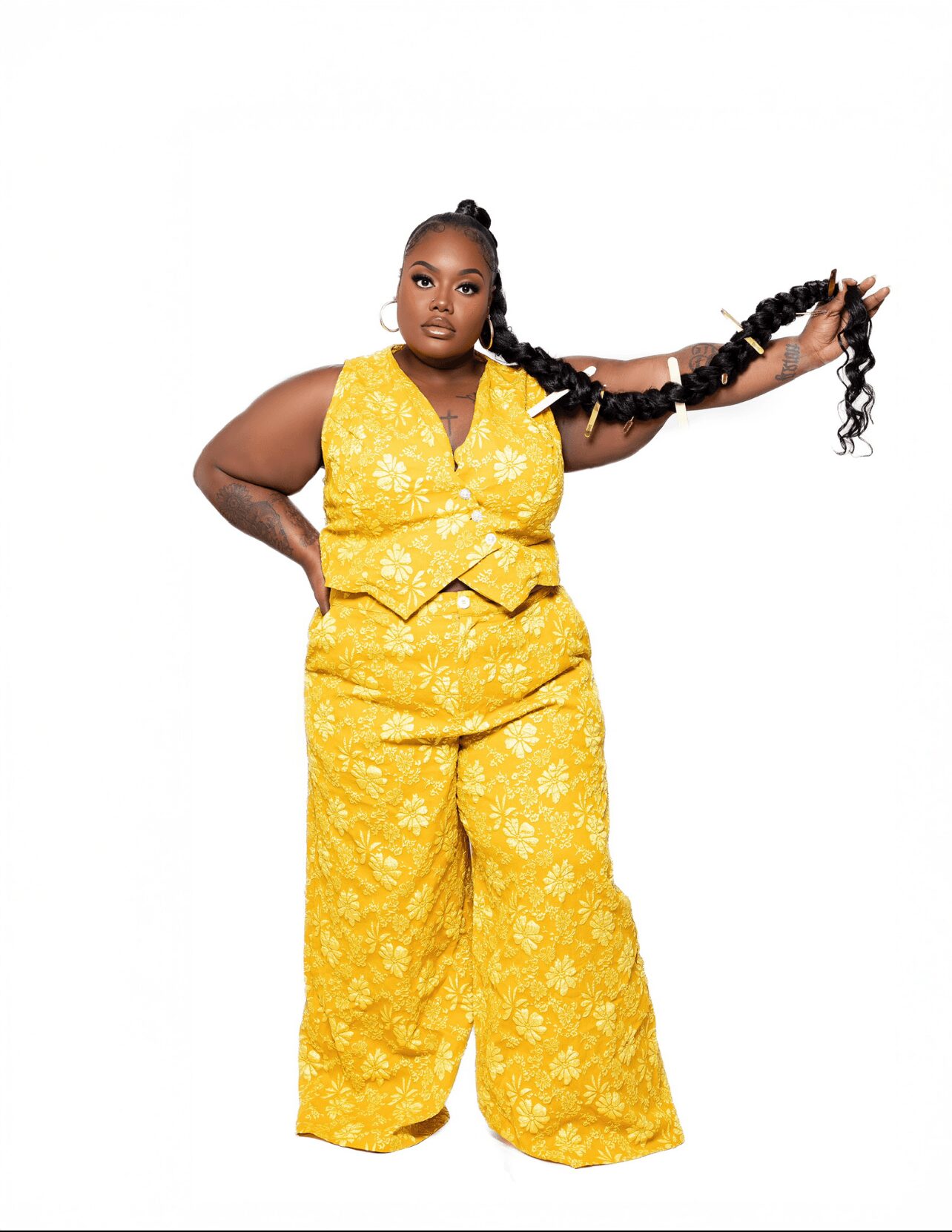 Woman in yellow patterned outfit holding long braided hair, standing against white background.