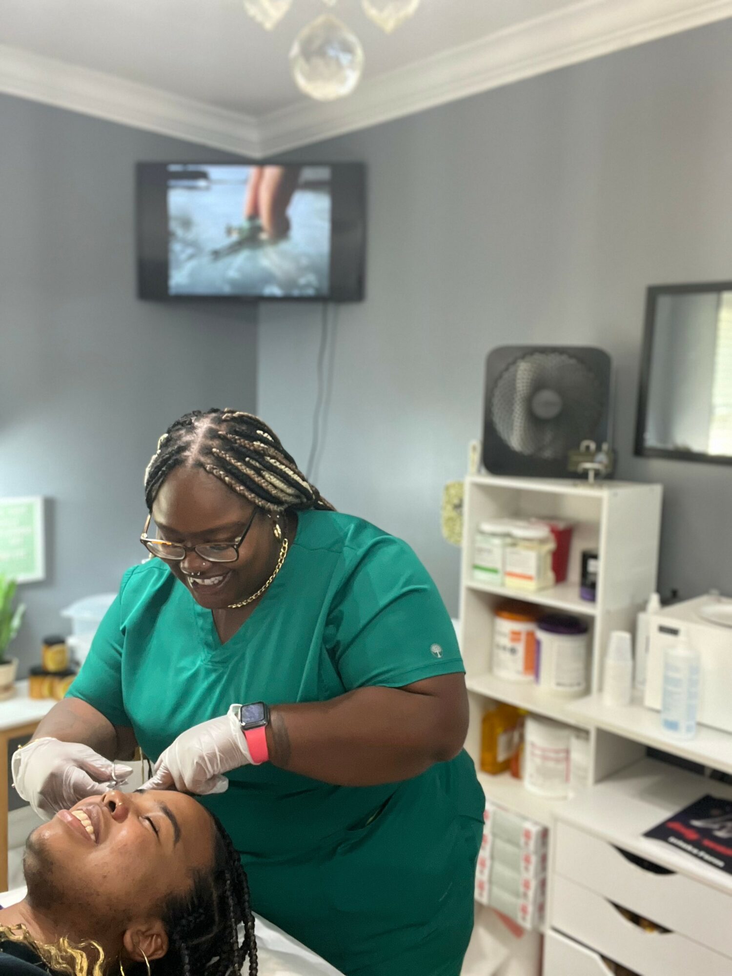 Healthcare worker in green scrubs smiling while attending to a patient in a clinical setting.