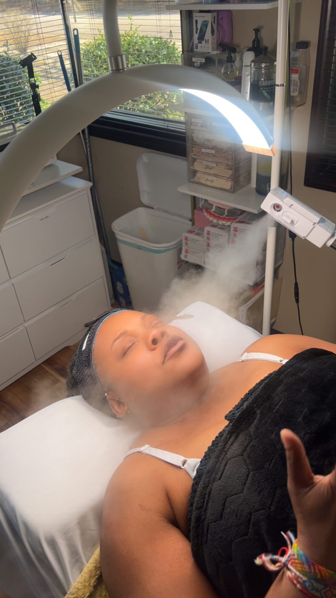 Woman lying on a treatment bed with a device emitting light and steam above her face in a clinical setting.