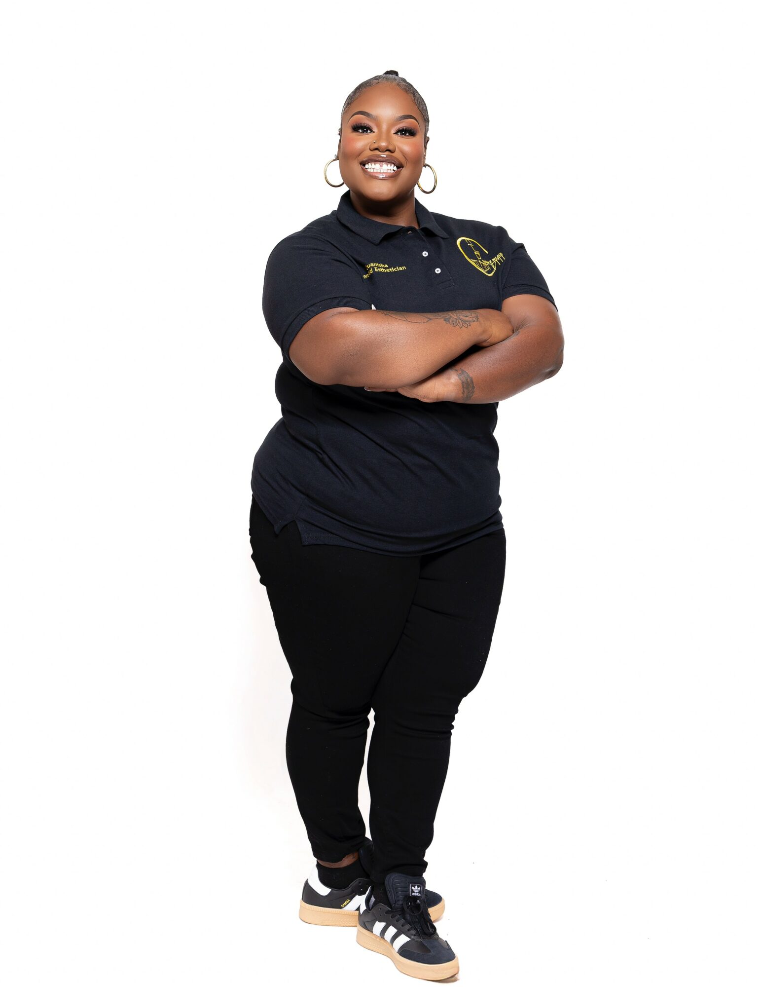 Person standing with arms crossed, smiling, wearing a black polo shirt and black pants, against a white background.