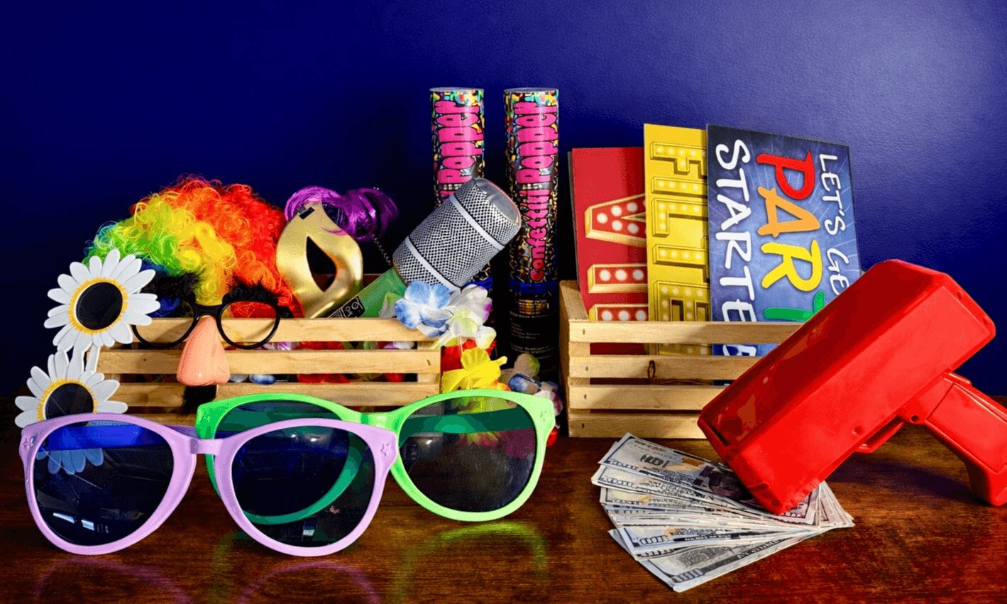 Colorful toys, sunglasses, a red cash register, and playing cards on a wooden surface against a blue background.