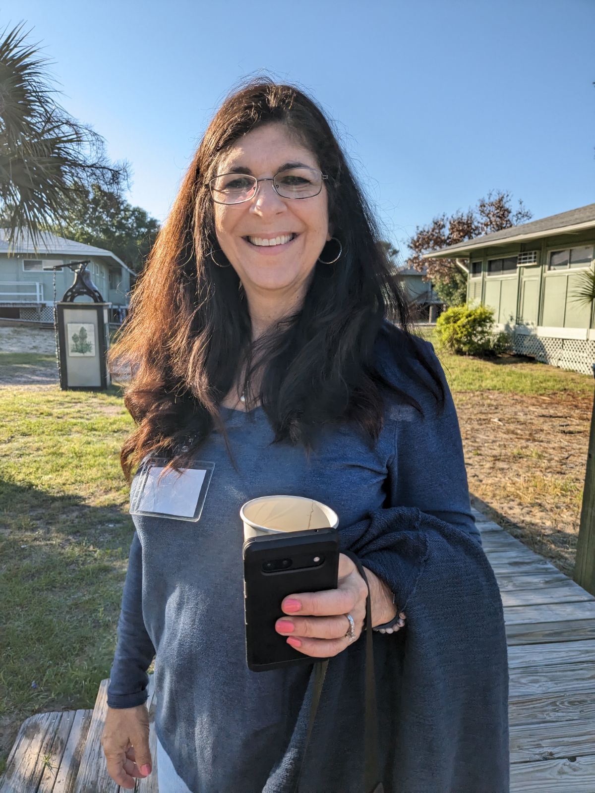 Woman with glasses smiling outdoors, holding a phone and a cup, in a park-like setting with trees and buildings.