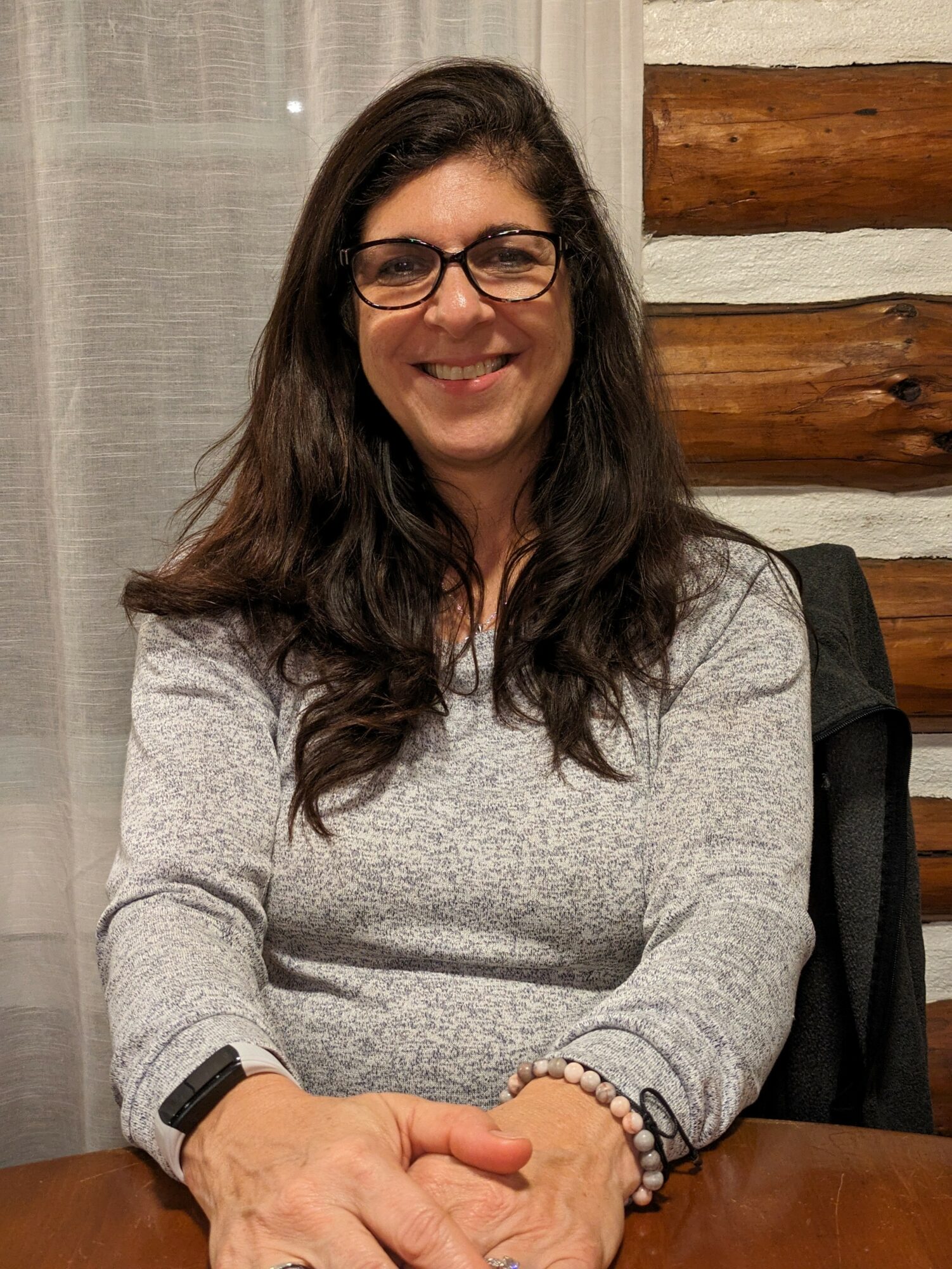 Woman with long dark hair and glasses smiling, sitting at a table indoors.