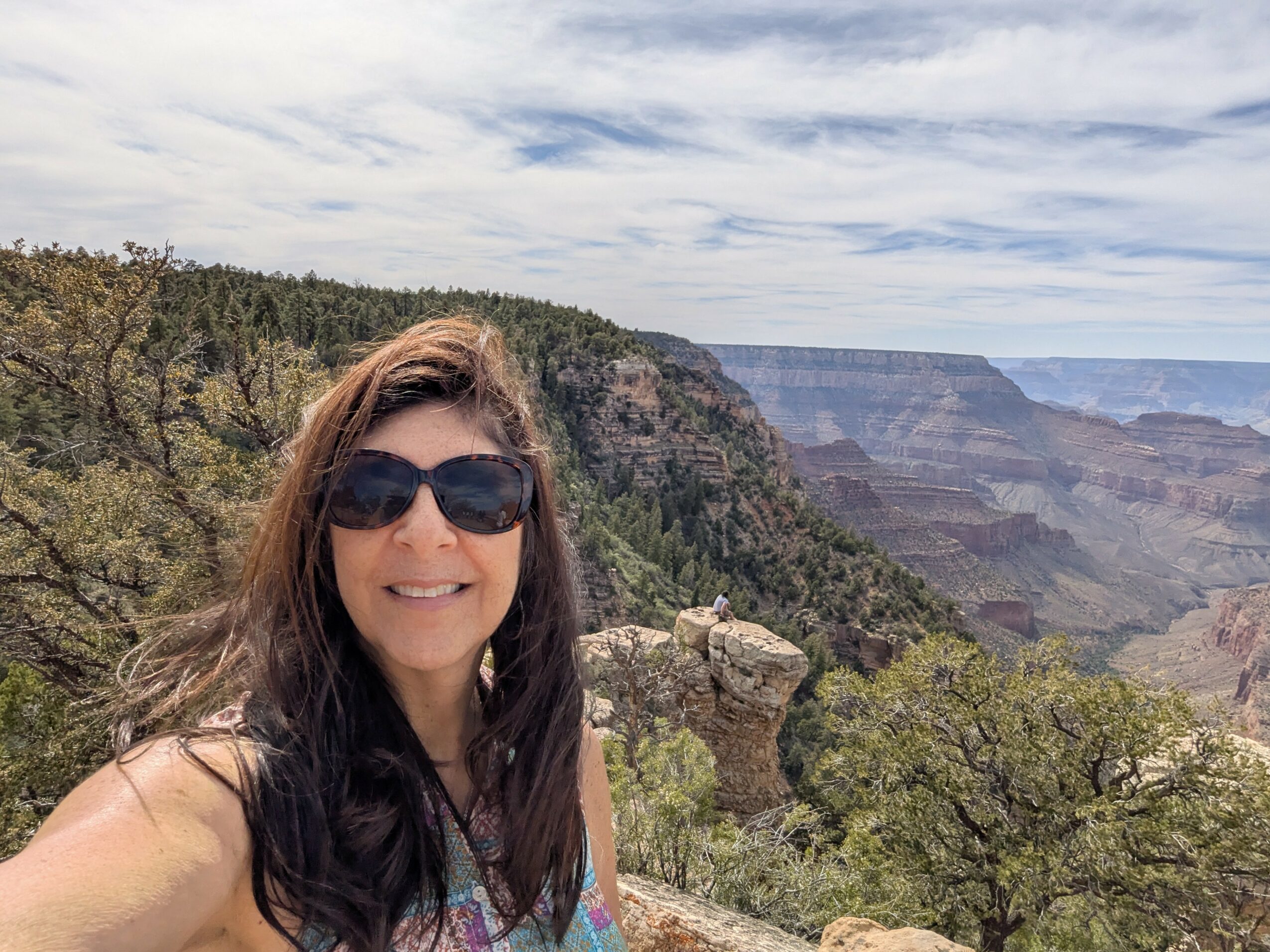 Woman with long dark hair and sunglasses smiling outdoors with canyon and trees in background.