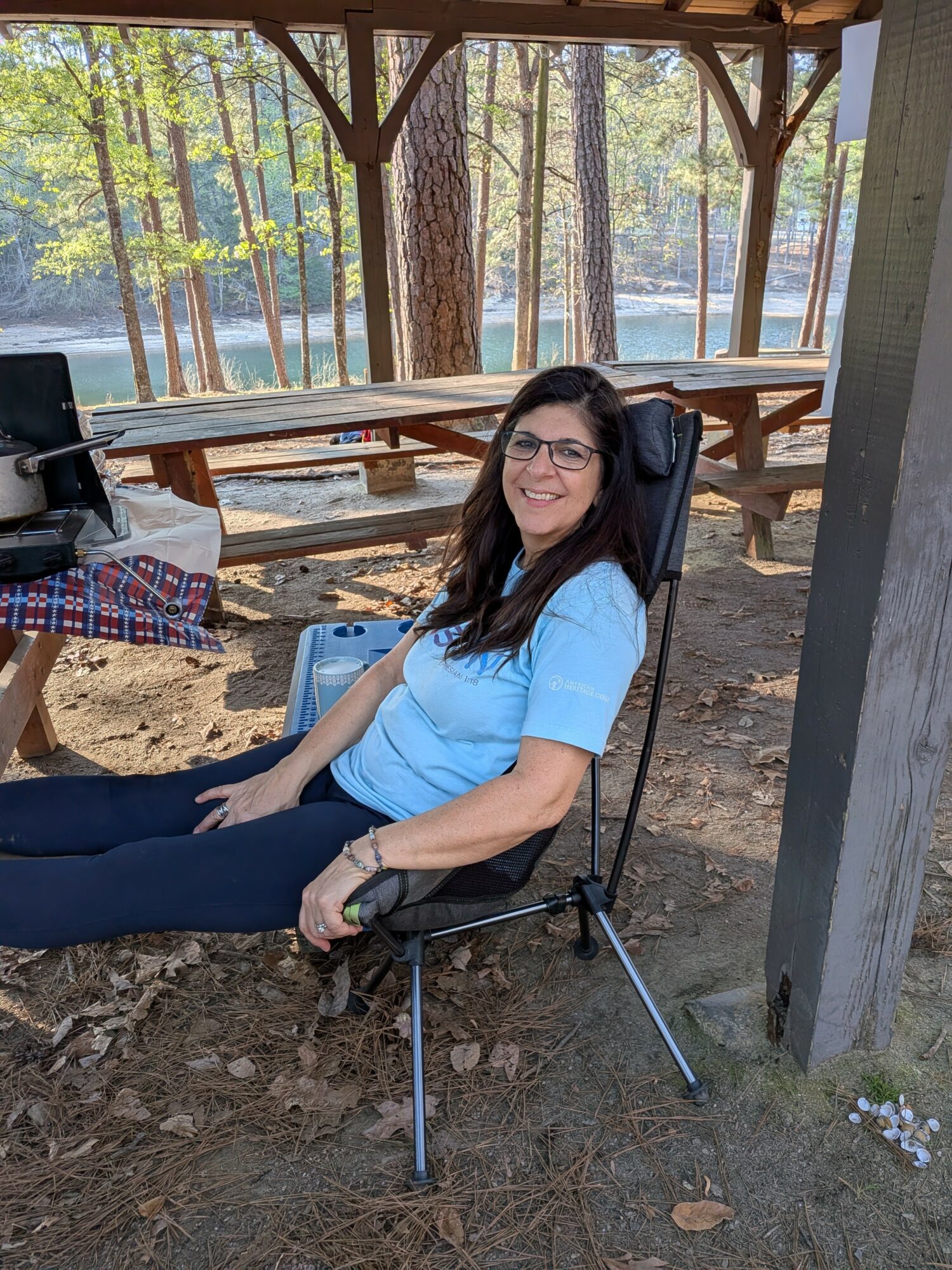 Woman sitting on a portable chair outdoors, smiling, wearing glasses and a light blue shirt, with trees in background.