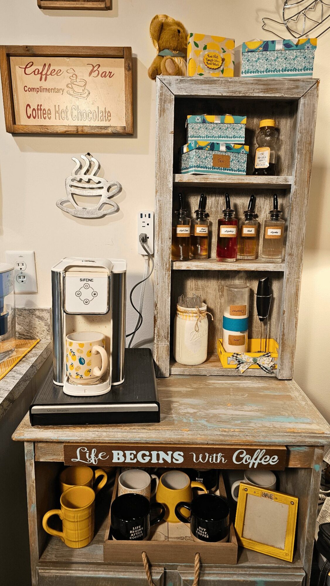 Coffee machine on table with yellow mugs, shelves with jars, a teddy bear, and decorative signs in background.