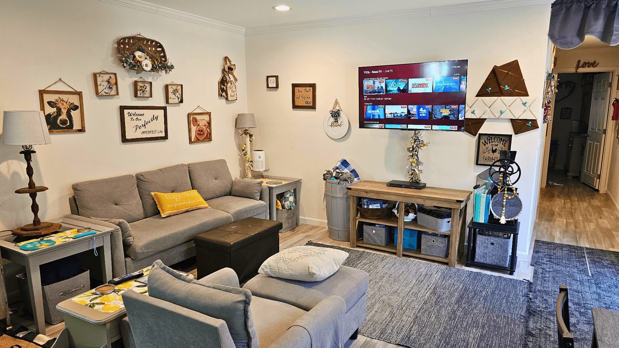 Living room with gray sofa, armchair, wall-mounted TV, framed pictures, and a wooden console table, with a hallway visible on the right.