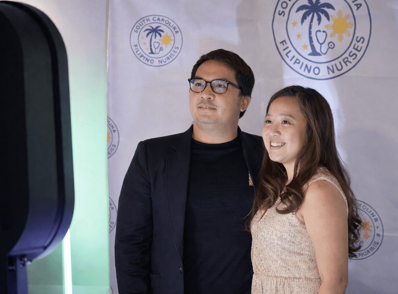 Man and girl stand together in front of a backdrop with logos, smiling and looking at a screen.