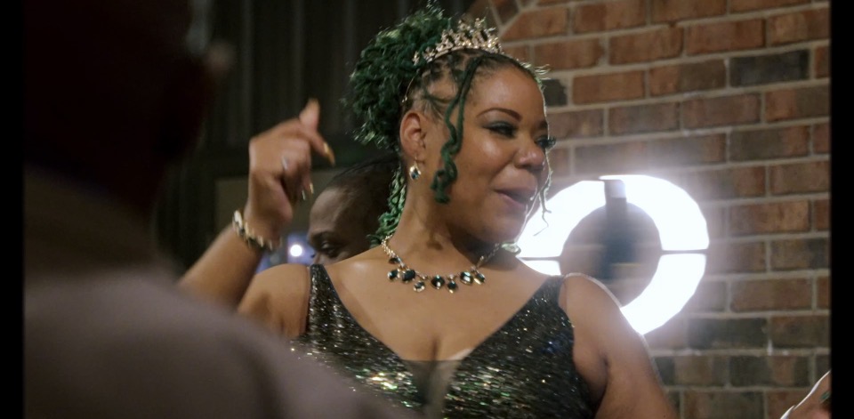 Woman with green hair and jewelry smiling, brick wall background, bright light behind her.
