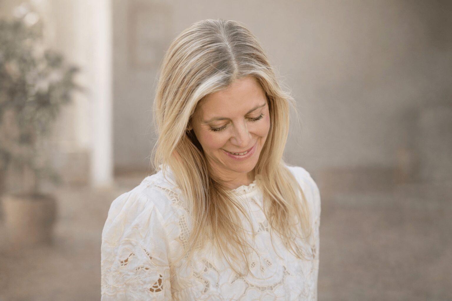 Woman with long blonde hair smiling, wearing a white lace top, in a softly lit indoor setting.