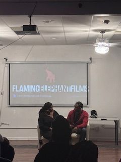 Two people sit and talk in front of a presentation screen in a room with a ceiling light and a table.