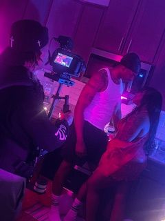 Three people in a dimly lit room with purple lighting, one standing and two sitting, with a camera on a tripod in the background.