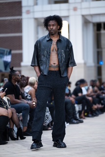 Person standing outdoors in front of seated crowd, wearing a denim jacket and dark pants.