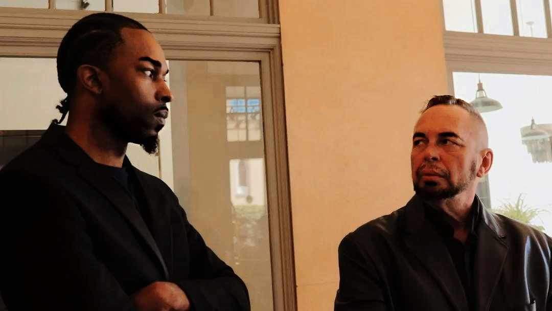 Two men engaged in conversation indoors, with large windows in the background and natural light illuminating the scene.
