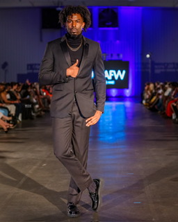 Male model walking on runway in a dark suit with a spotlight, audience seated on both sides, blue background.