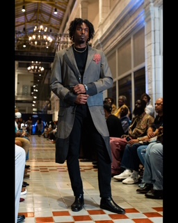 Man with curly hair in a gray blazer and black pants walking in a large, ornate hall with seated audience.