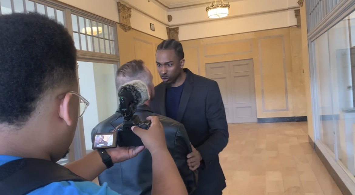 Three people in a room, one holding a camera, two facing each other, with large windows and wooden floors.