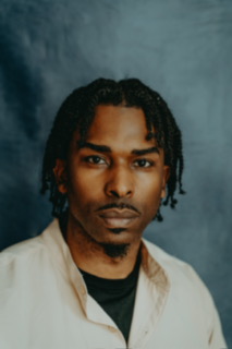Portrait of a man with dark hair styled in twists, wearing a light-colored shirt, against a textured background.