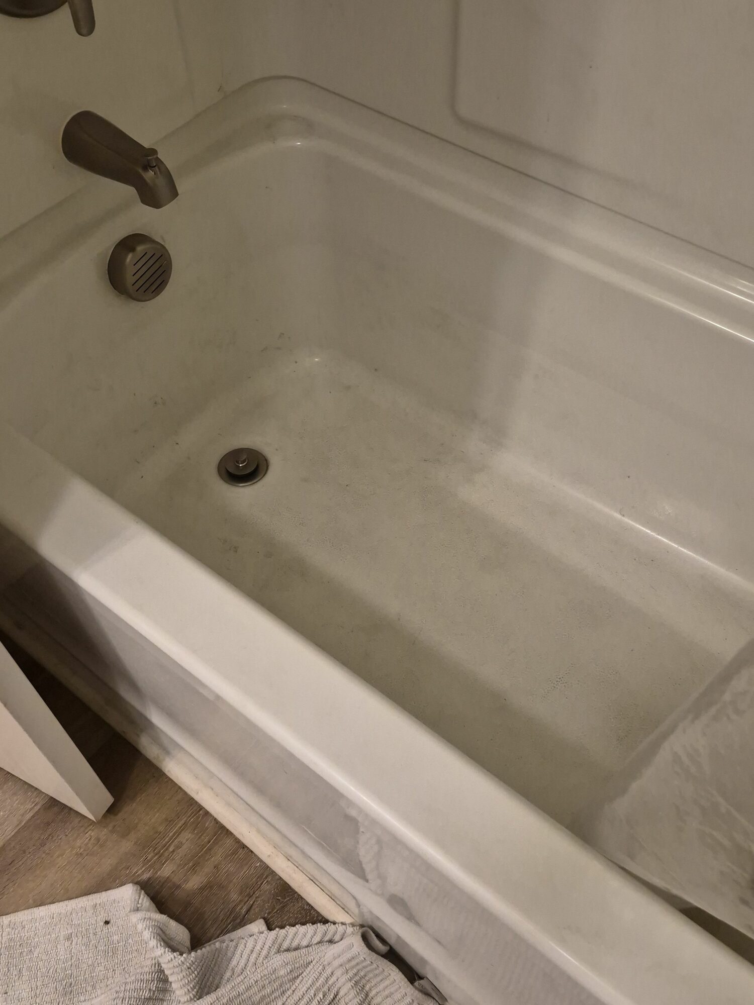 Empty bathtub with drain and faucet, surrounded by white walls and a wooden floor mat.