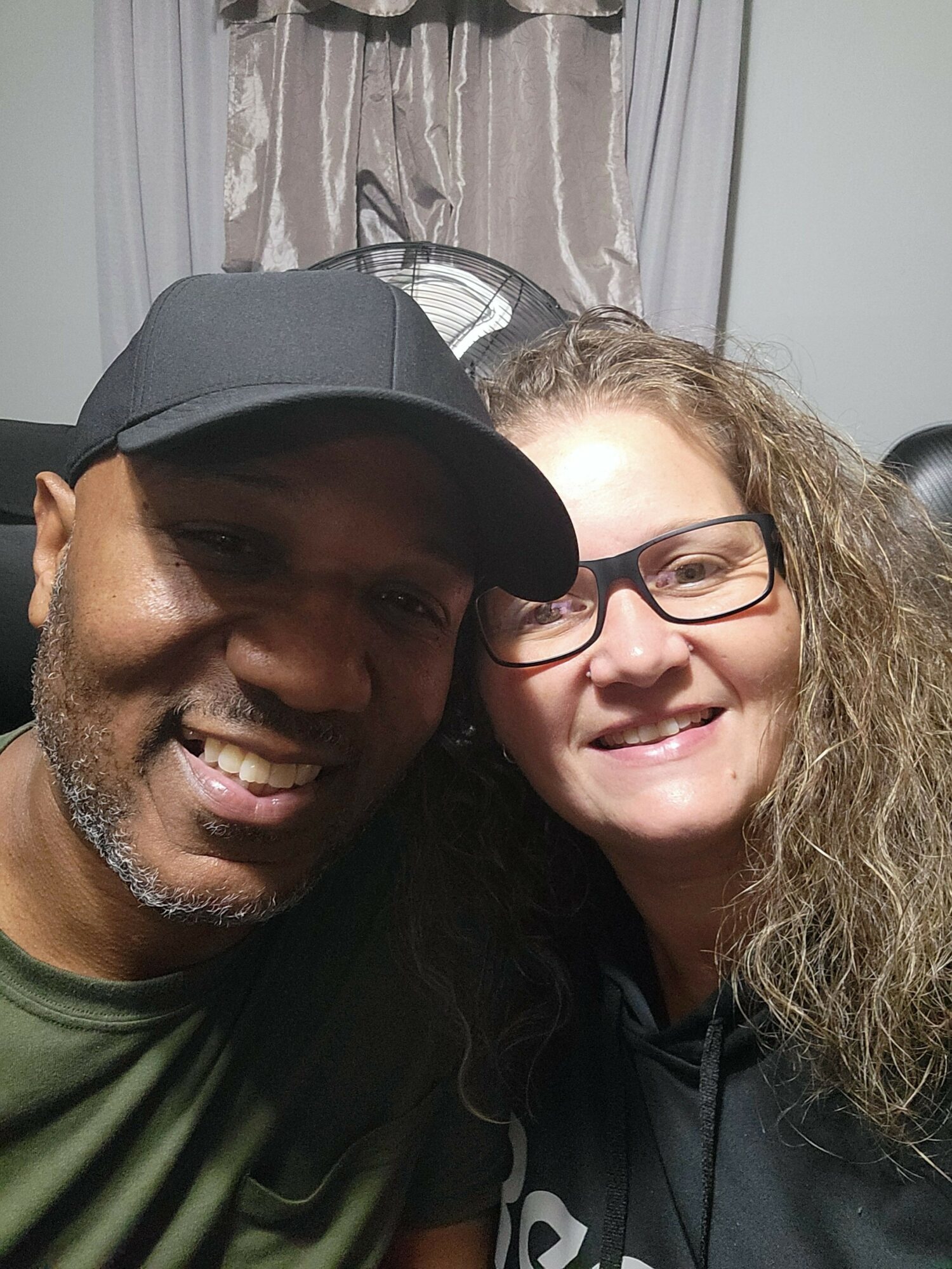 Two smiling people close together, one wearing a black cap and the other with glasses and curly hair.