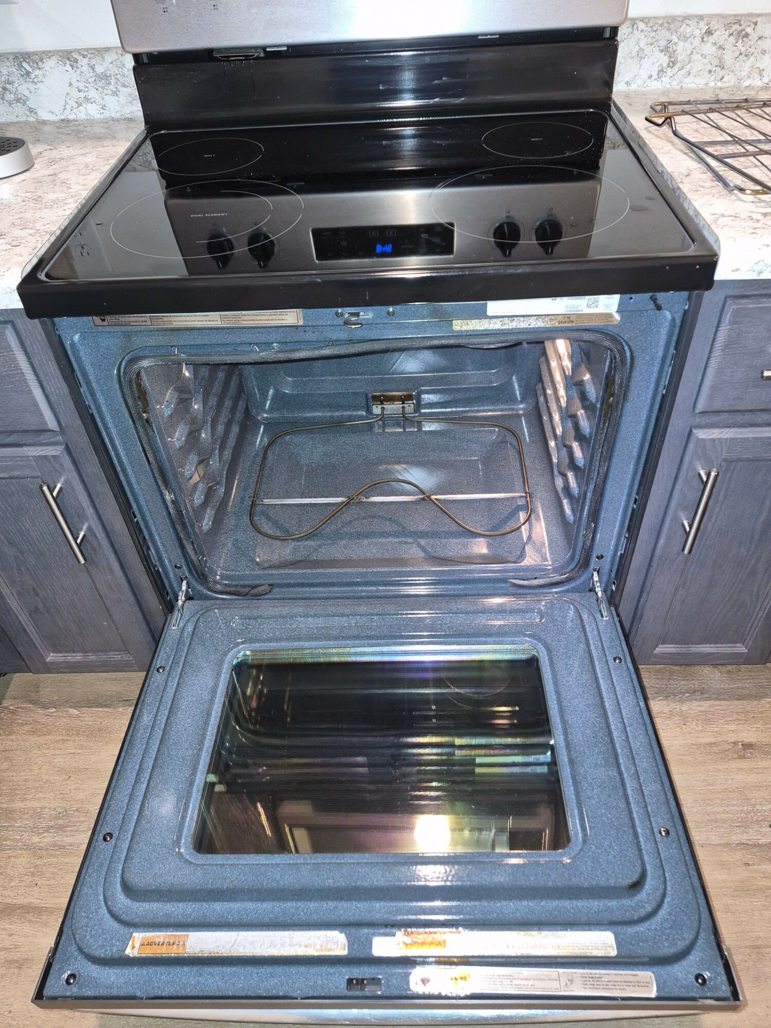 Open oven door revealing empty oven interior with glass window and control panel above, stove on top, kitchen cabinets on sides.
