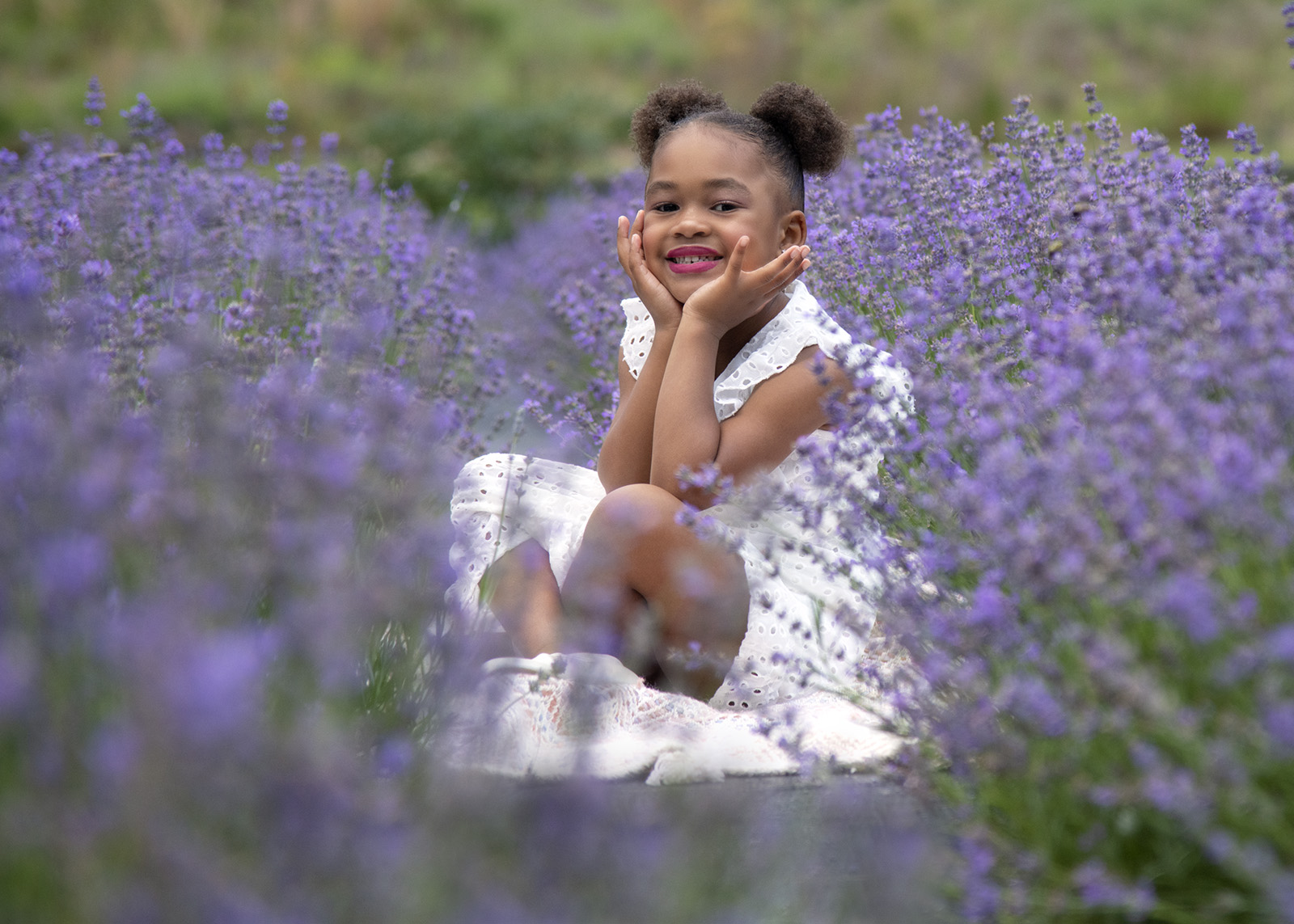 Young girl sitting among purple flowers, smiling with hands on cheeks, wearing a white dress.