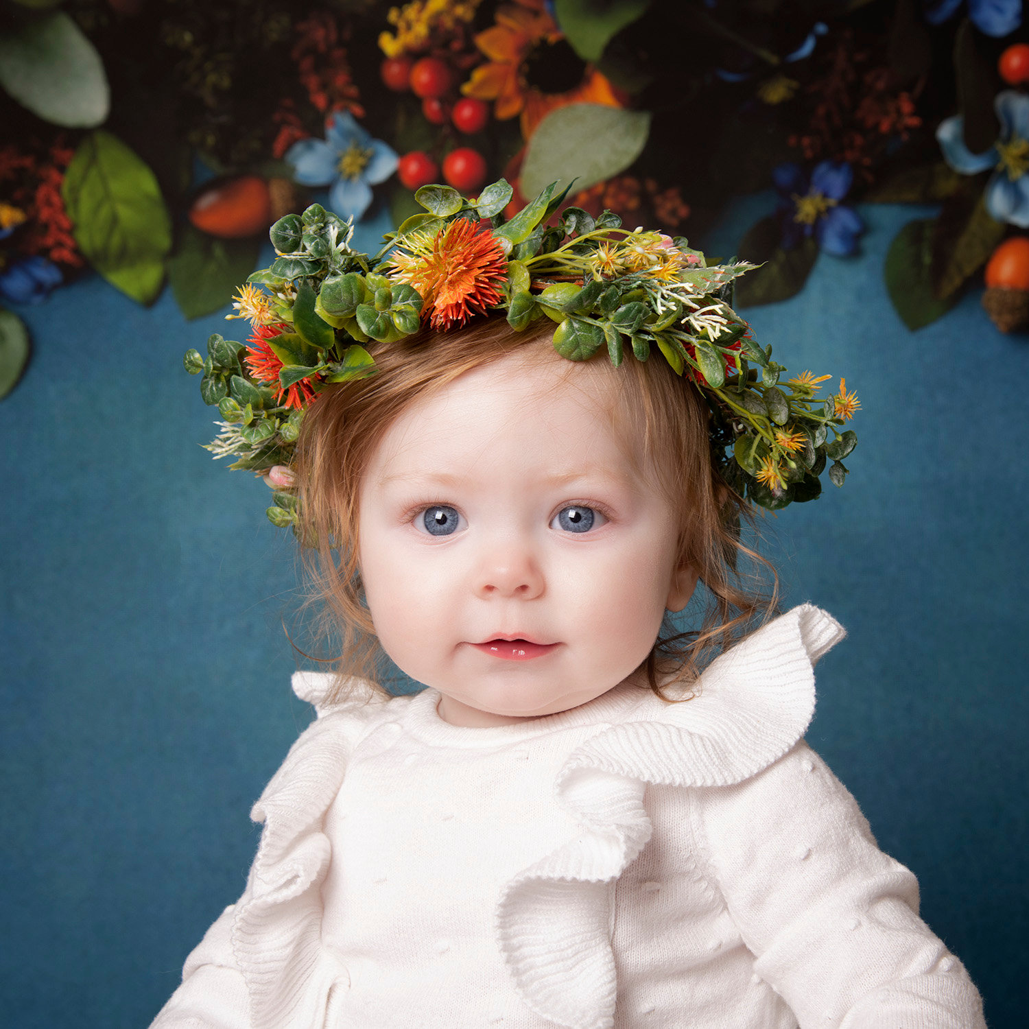 Child with a floral crown, wearing a white ruffled dress, against a colorful floral background.