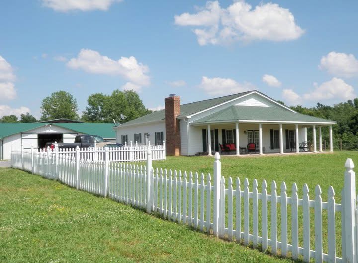 White house with porch, brick chimney, surrounded by a white picket fence, green lawn, blue sky with clouds.