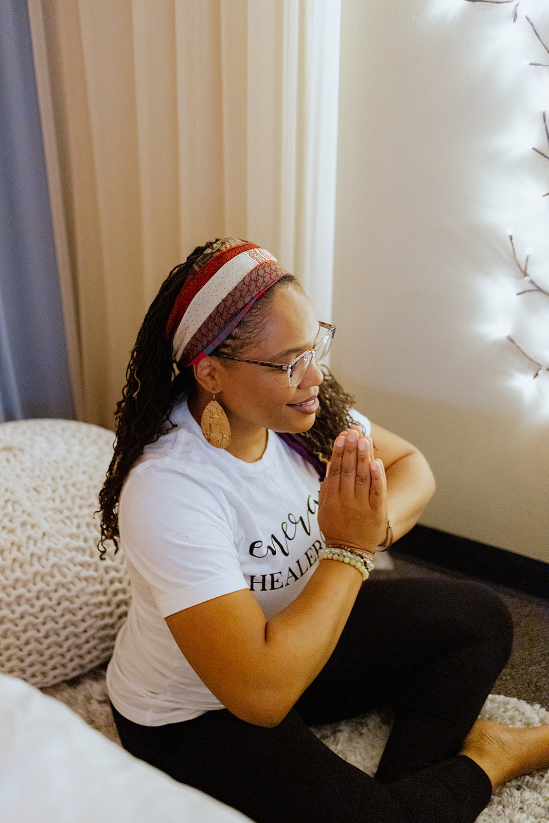 Woman sitting on a couch with hands in prayer position, wearing glasses, a headband, and a white t-shirt.