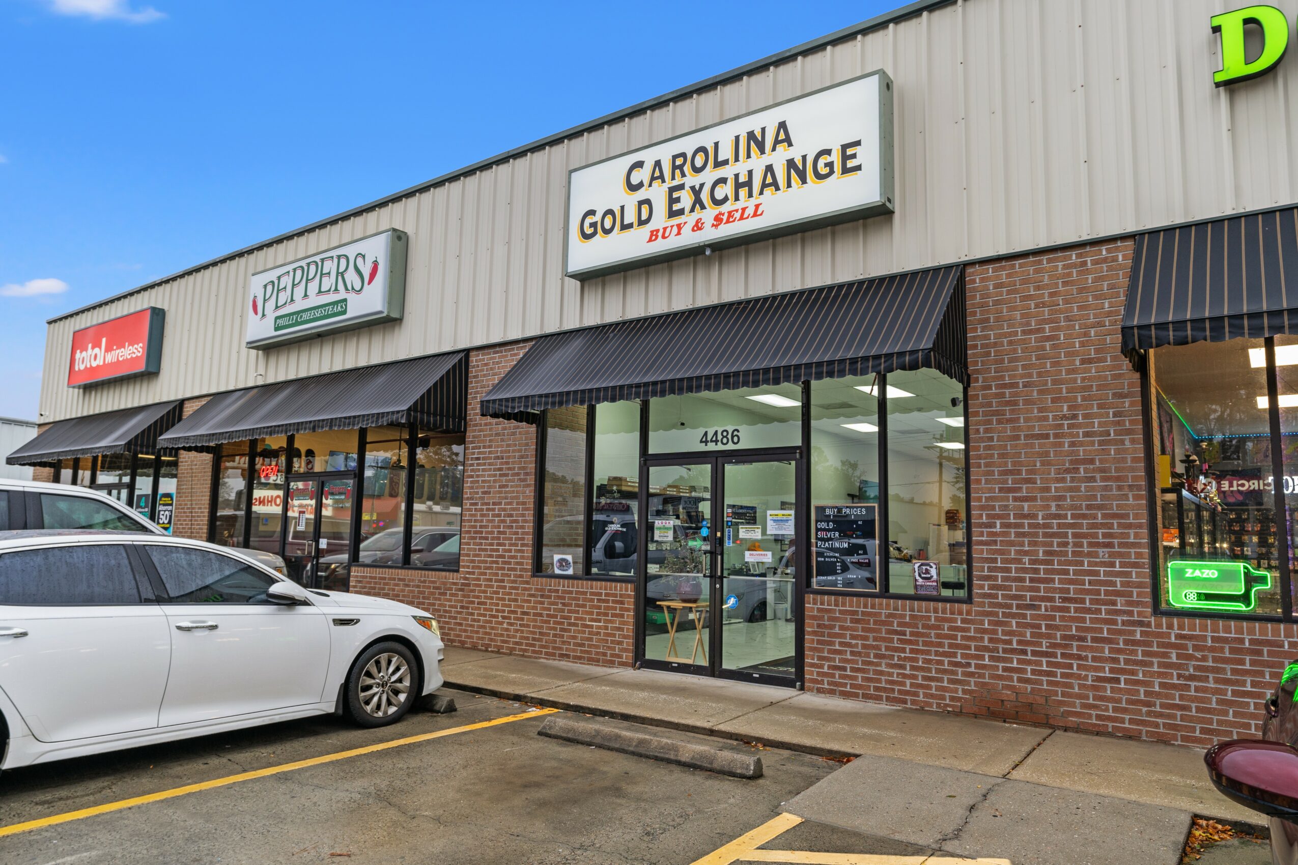 Strip mall storefronts with signs for Peppers and Carolina Gold Exchange, parked car in front, blue sky above.