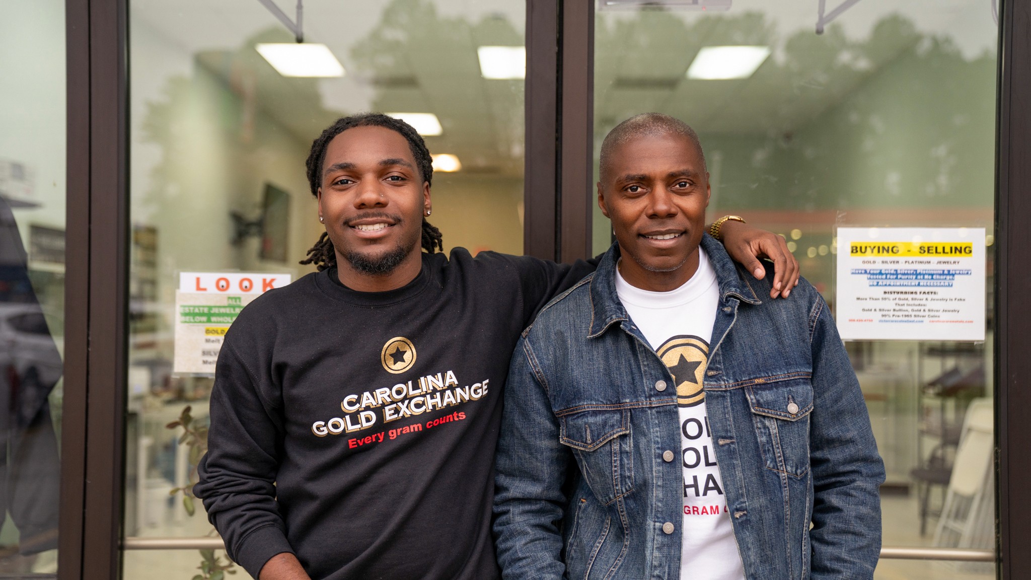Two men standing outside a glass door, smiling, with one arm around each other's shoulders.