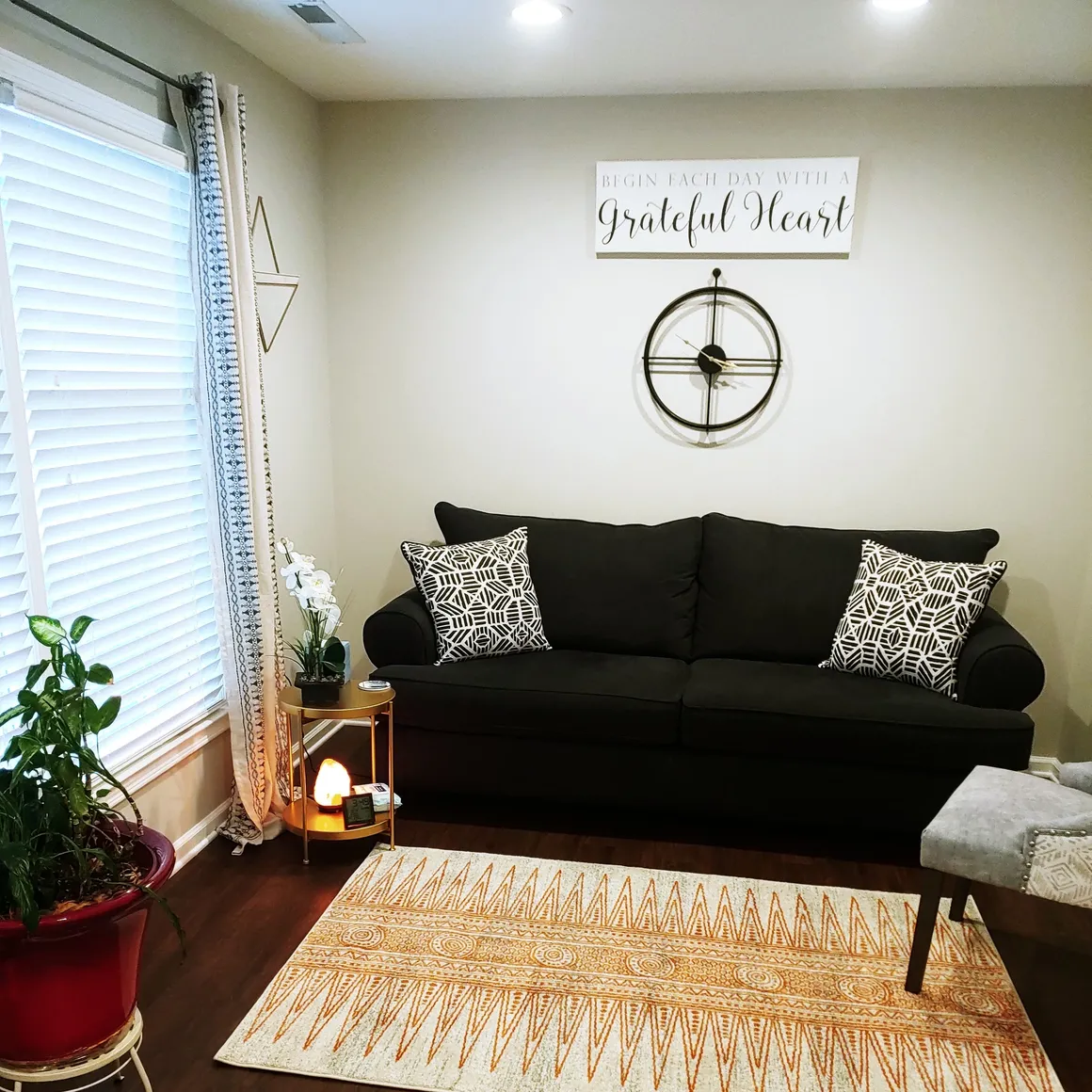 Living room with black sofa, patterned pillows, wall clock, wall sign, window with blinds, potted plant, and patterned rug.