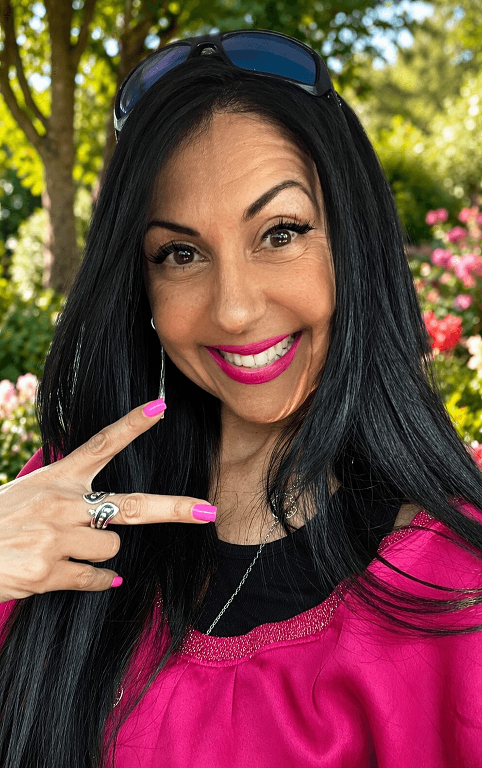 Woman with long black hair smiling outdoors, wearing sunglasses on head and pink top, pointing at her face.
