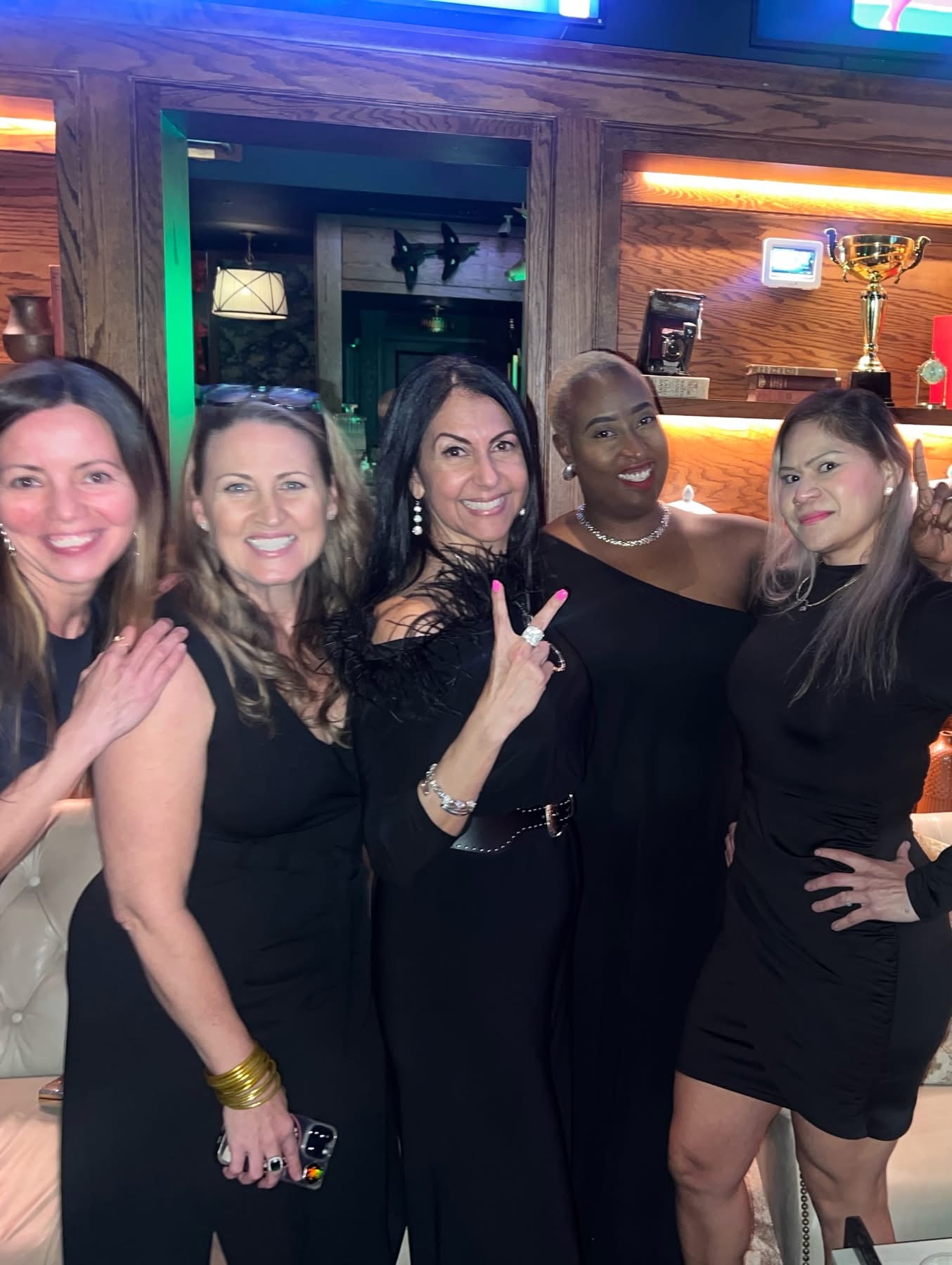 Four women standing together indoors, smiling, with one making a peace sign, in a bar or restaurant setting.