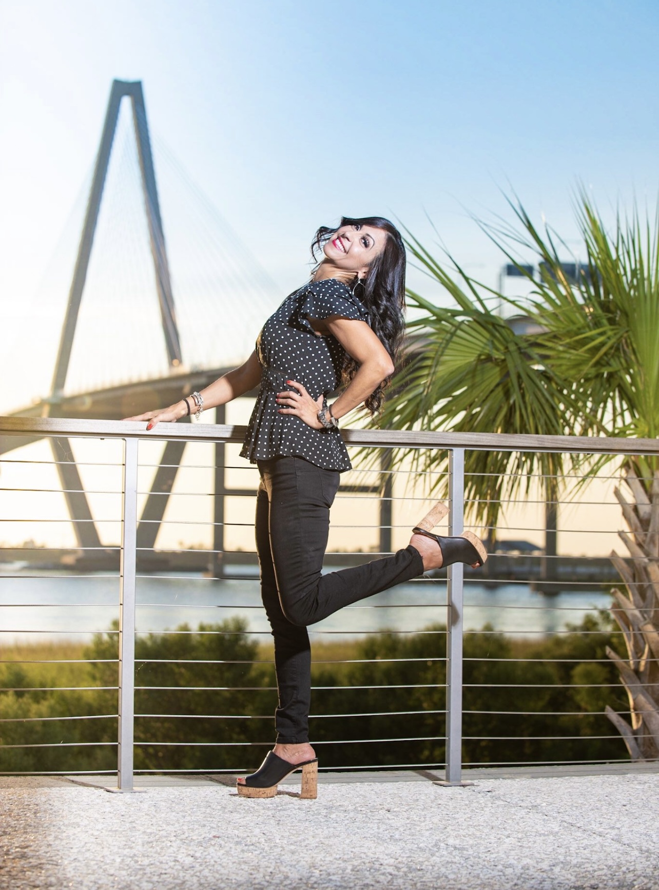 Woman standing on one leg, smiling, outdoors with a bridge and palm tree in background.