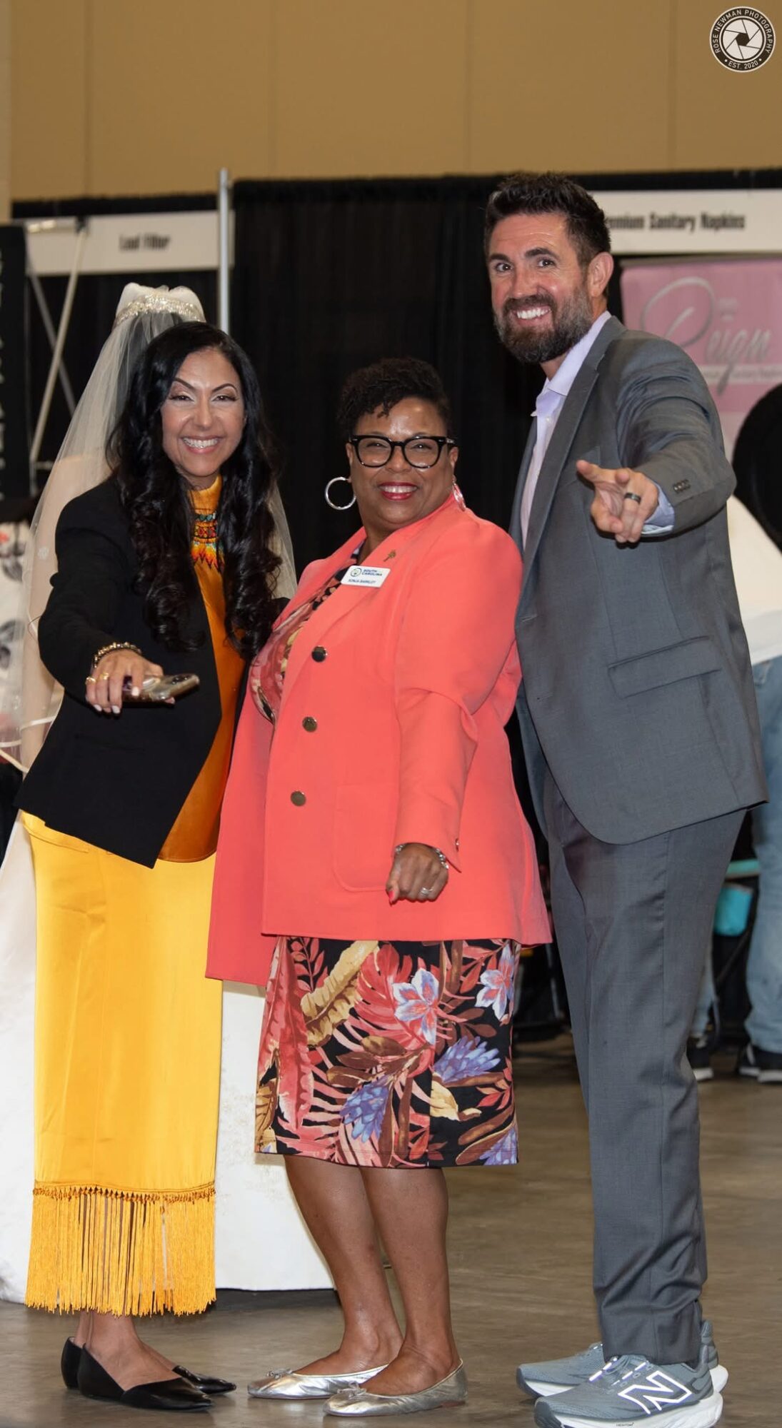 Three people standing together, smiling, indoors with booths in background.