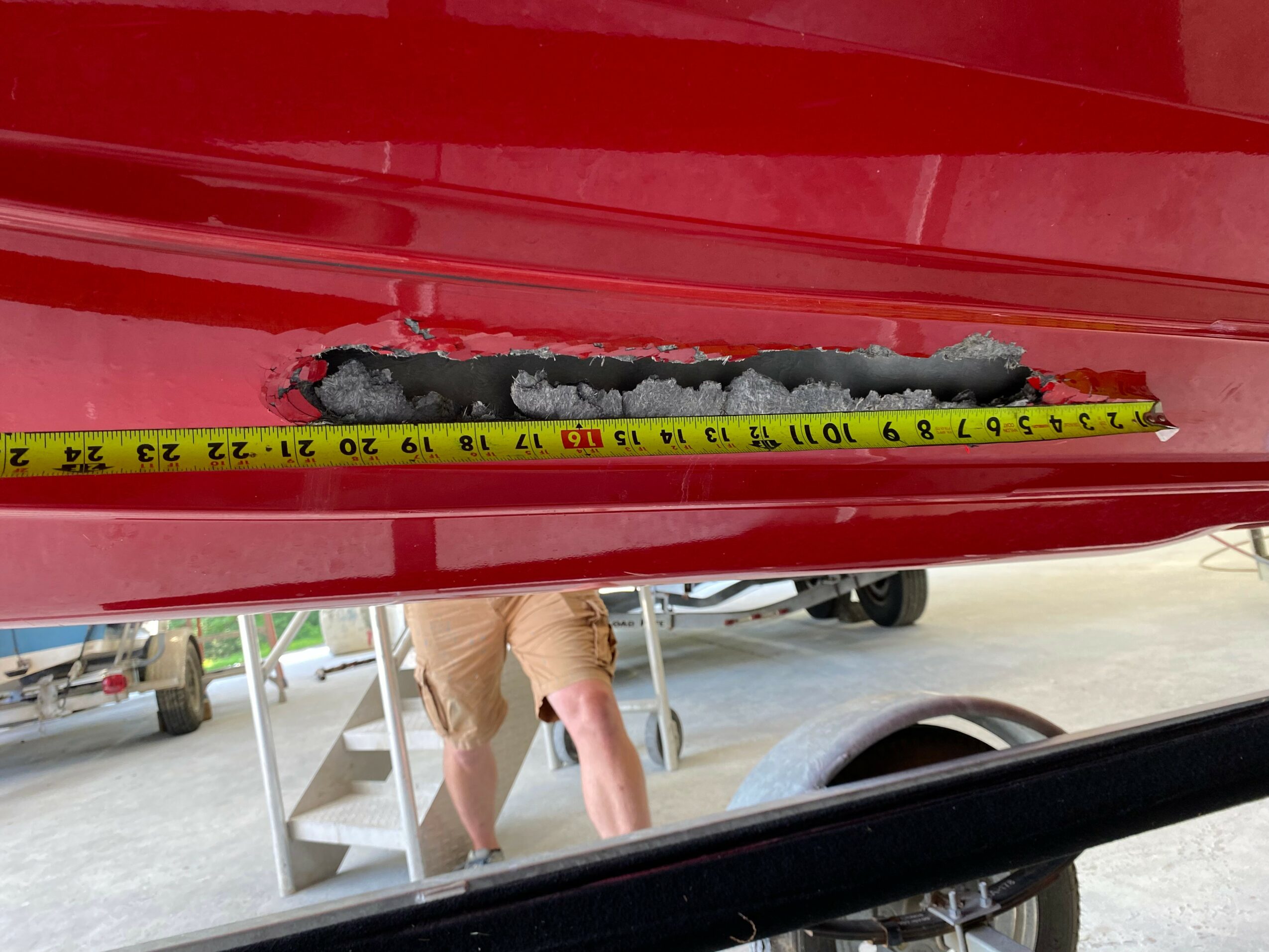 Close-up of a damaged red surface with a measuring tape showing length, and a person in shorts working nearby.
