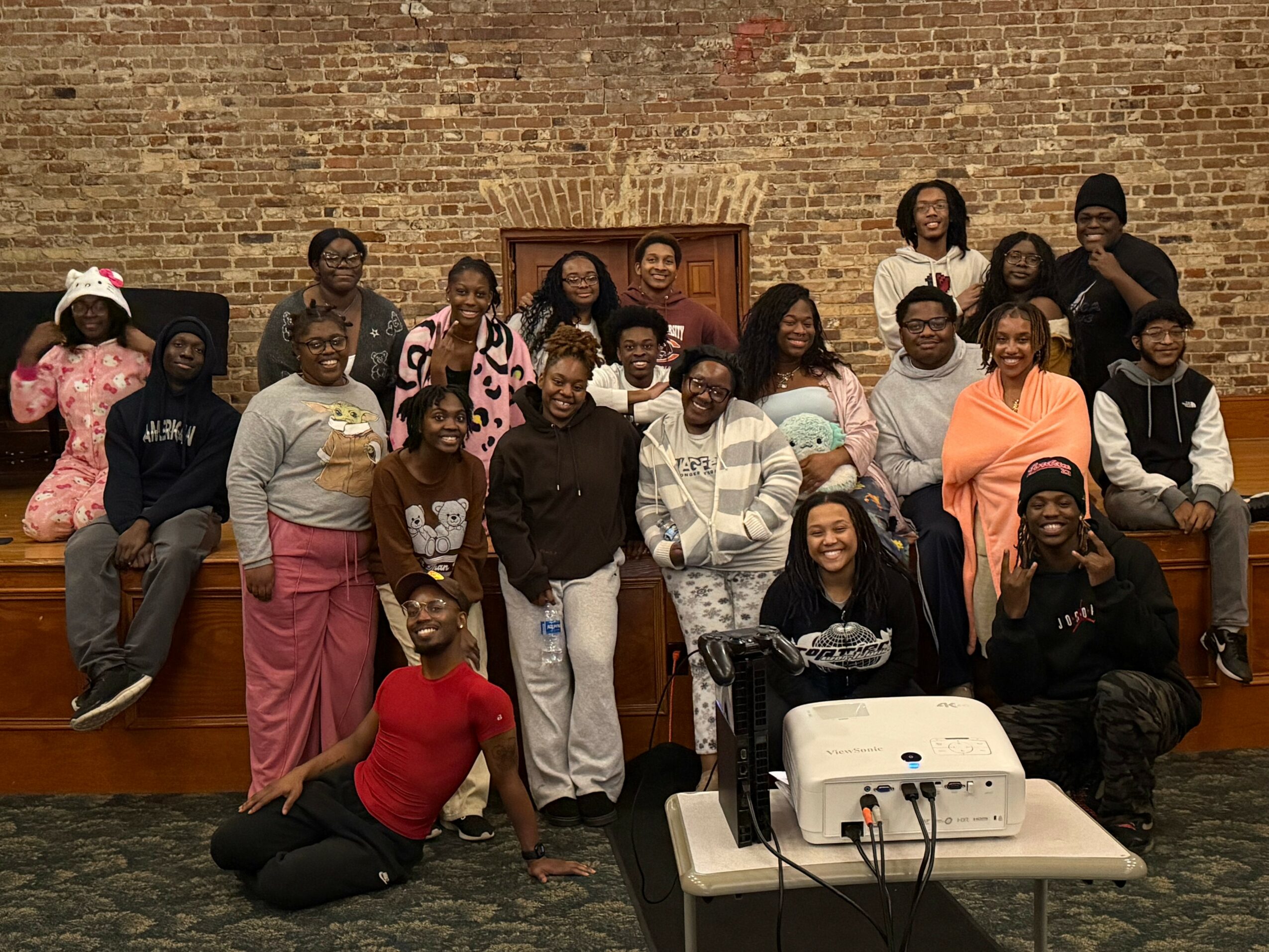 Group of people posing indoors against a brick wall, some sitting and some standing, with a projector on a table.