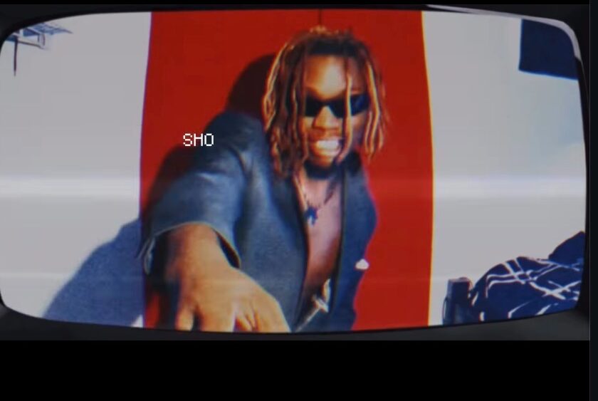 Person with dreadlocks and sunglasses leaning forward, with a red and white background.