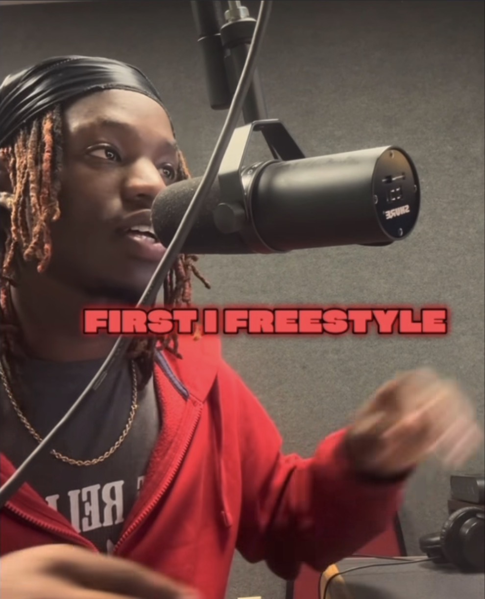 Person with dreadlocks speaking into a microphone in a recording studio, wearing a red hoodie.