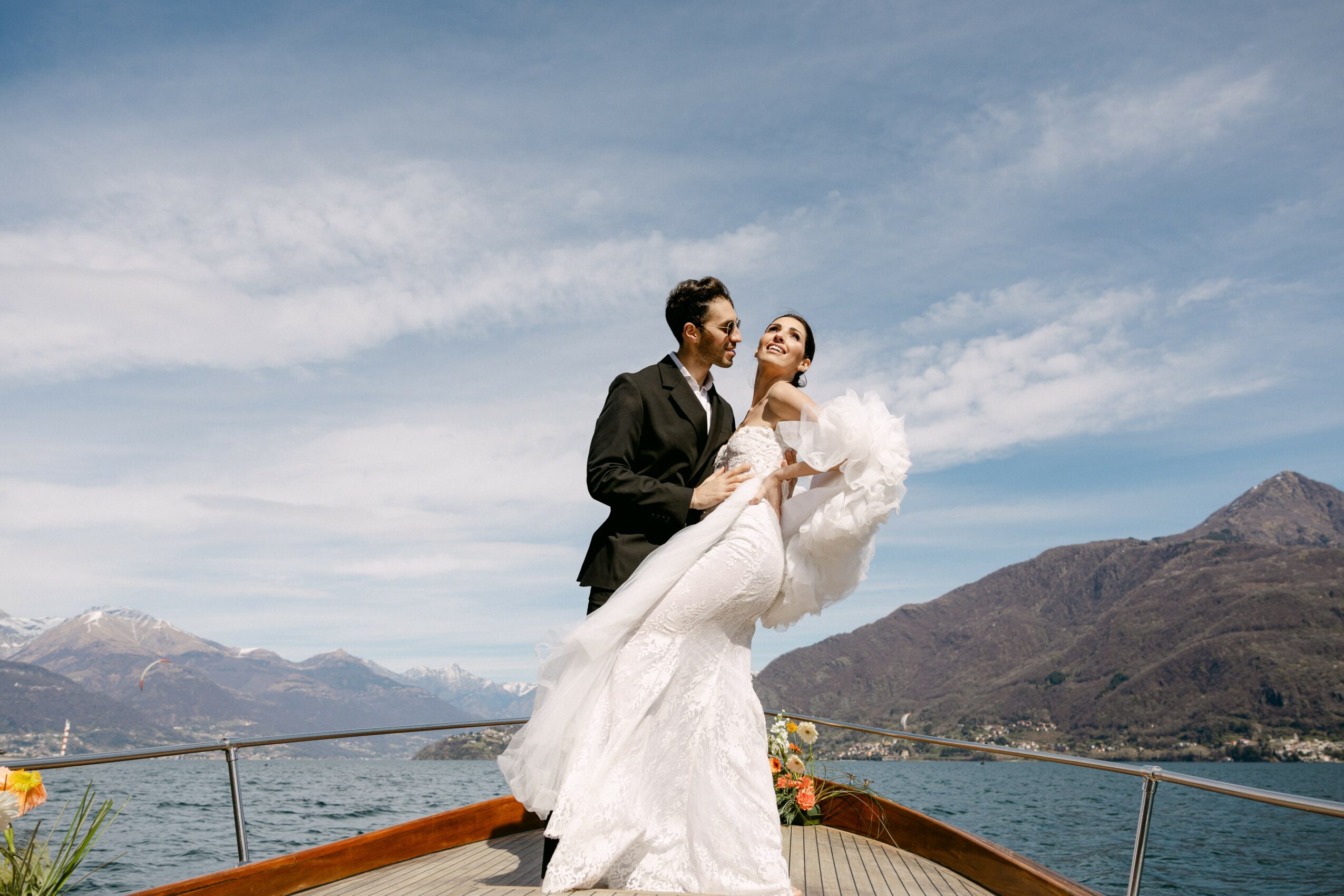 A bride and groom on a boat with mountains and sky in the background, the groom lifting the bride in a wedding dress.