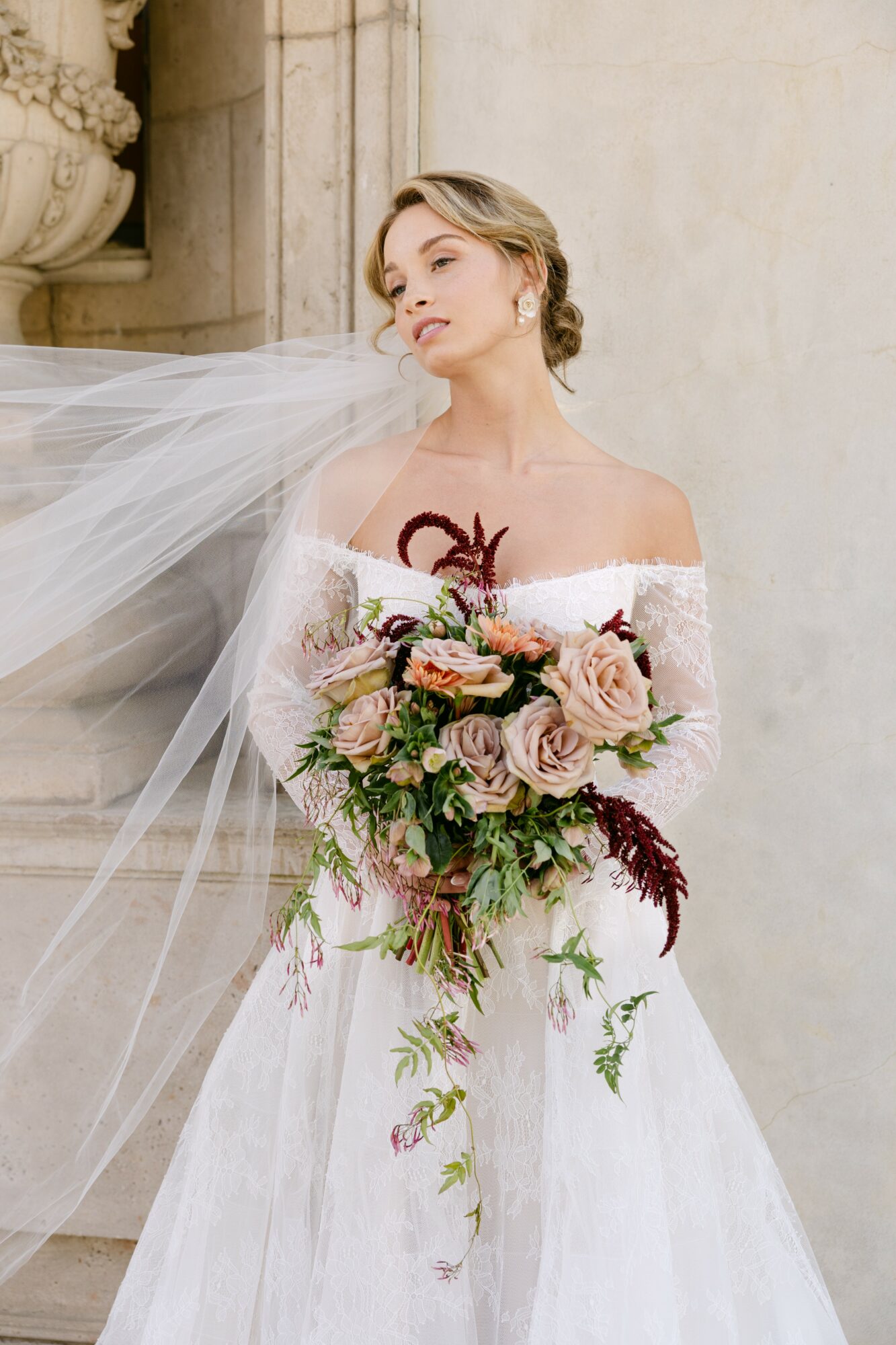 Woman in wedding dress holding a large bouquet of pink and purple flowers, standing outdoors near stone architecture.
