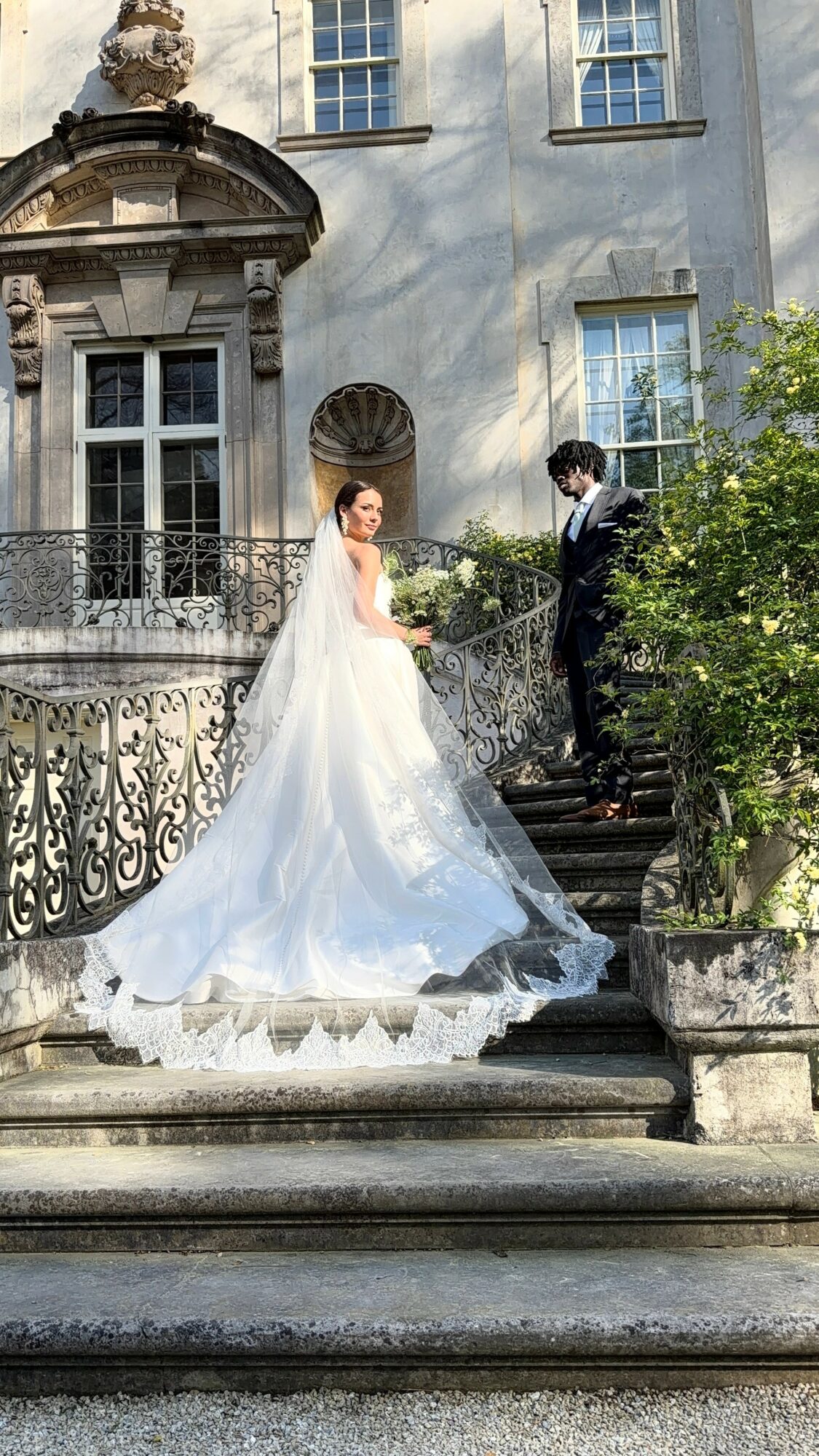 Bride and groom on stairs outside a building, bride in wedding dress, groom in suit, greenery nearby.