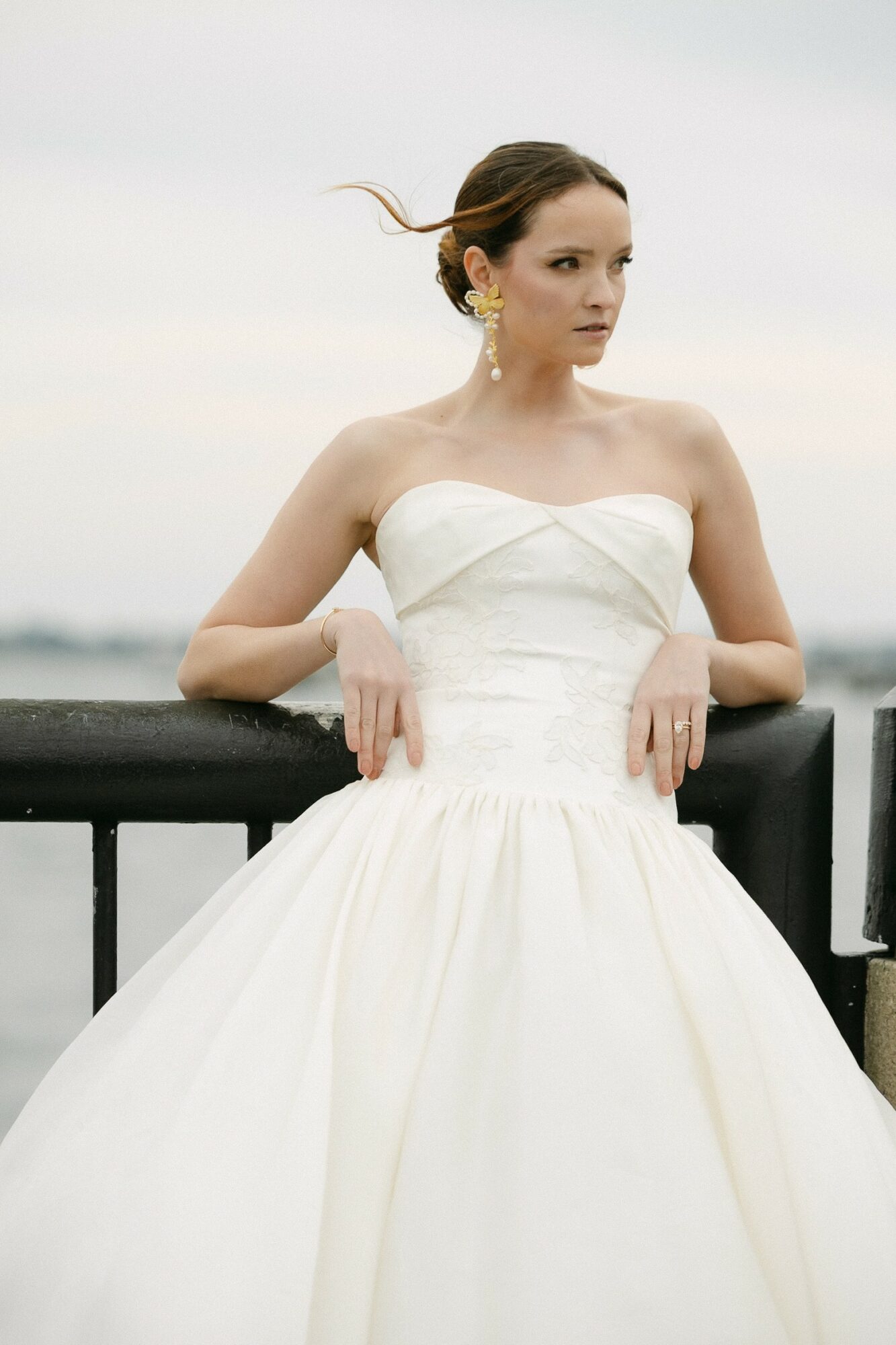 Woman in white strapless dress leaning on a black railing outdoors, with cloudy sky background.