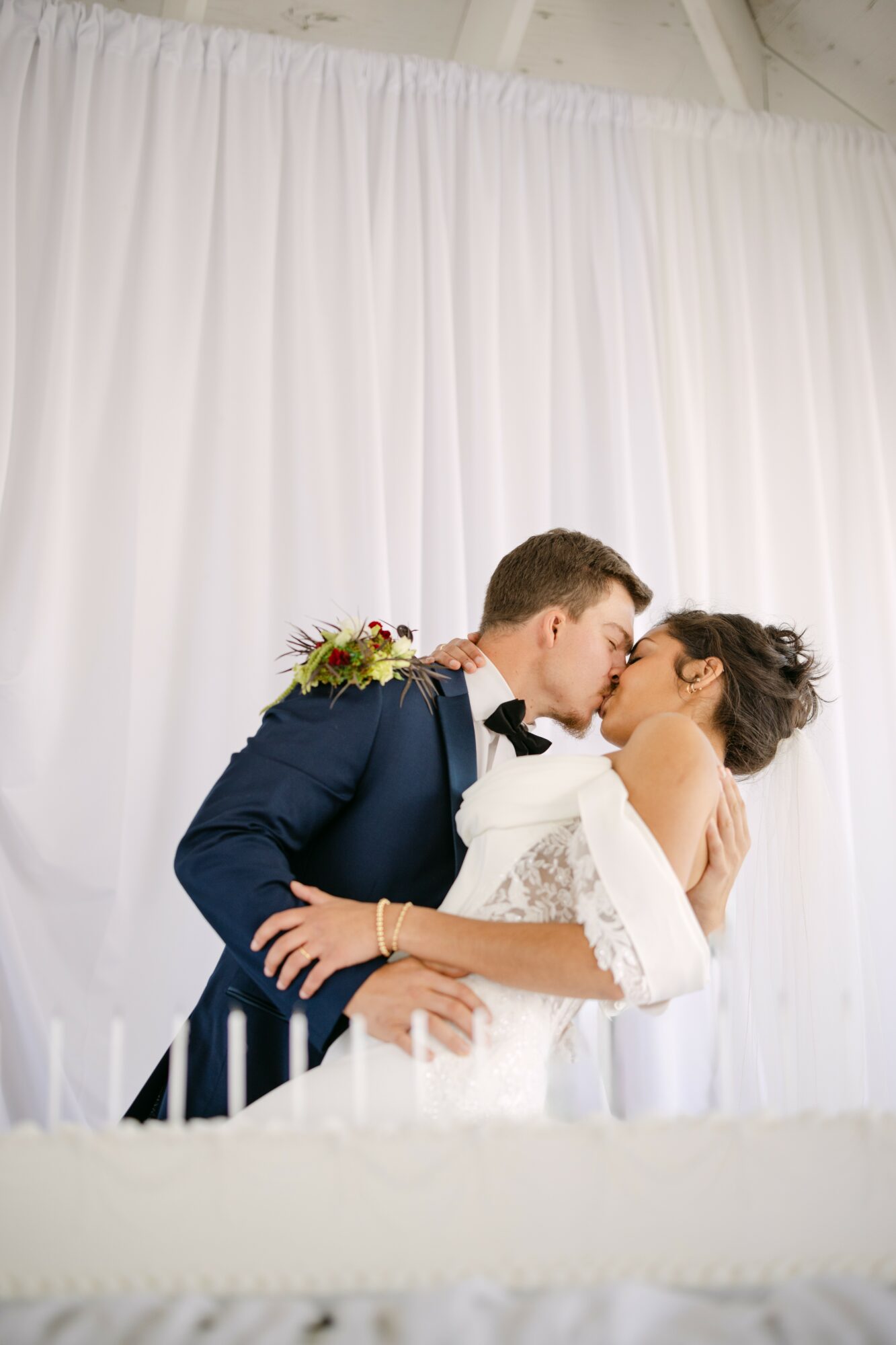Couple in wedding attire sharing a kiss in front of a white curtain backdrop.