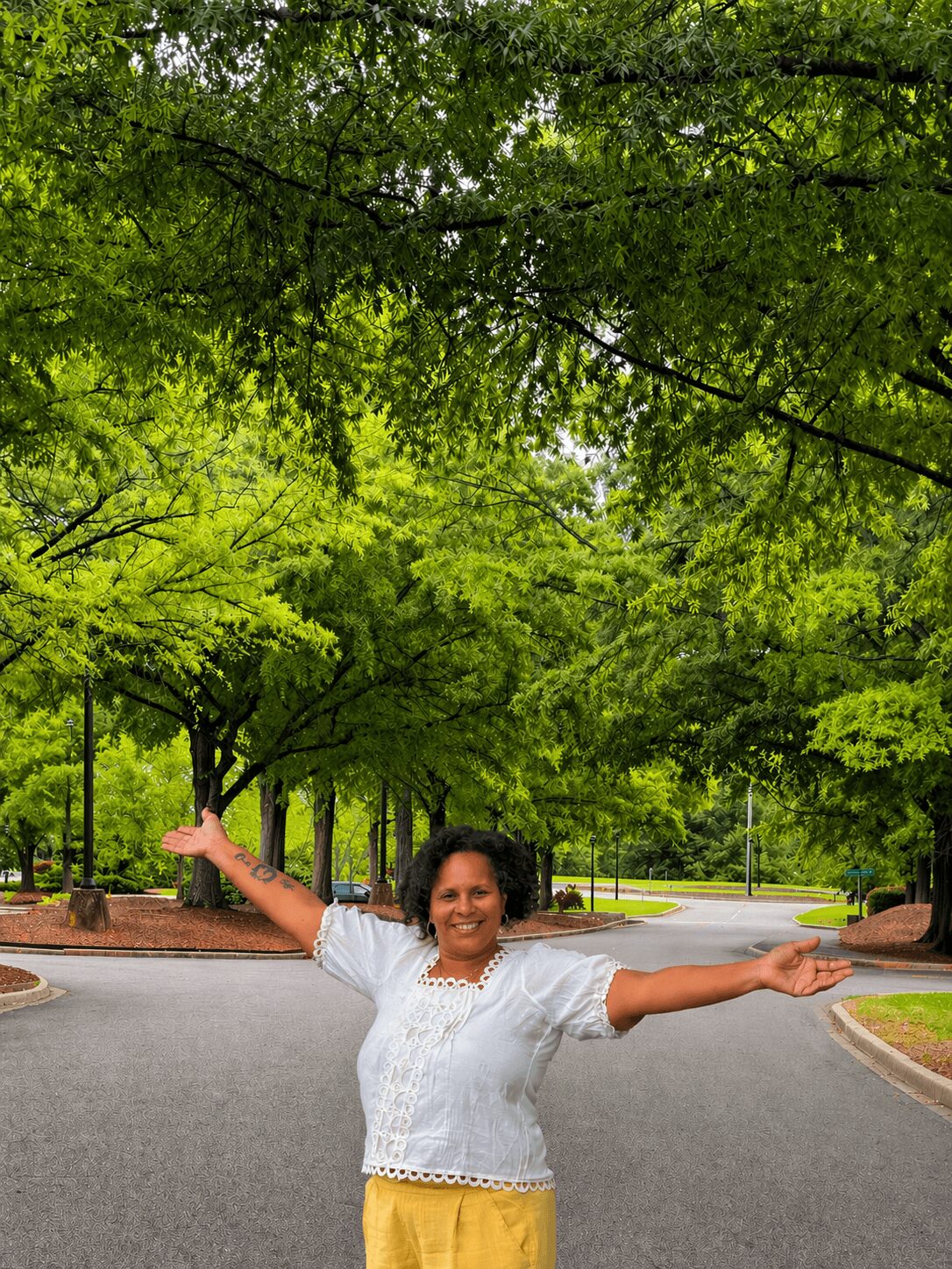 Woman standing on a street with arms outstretched, surrounded by green trees and a clear sky.
