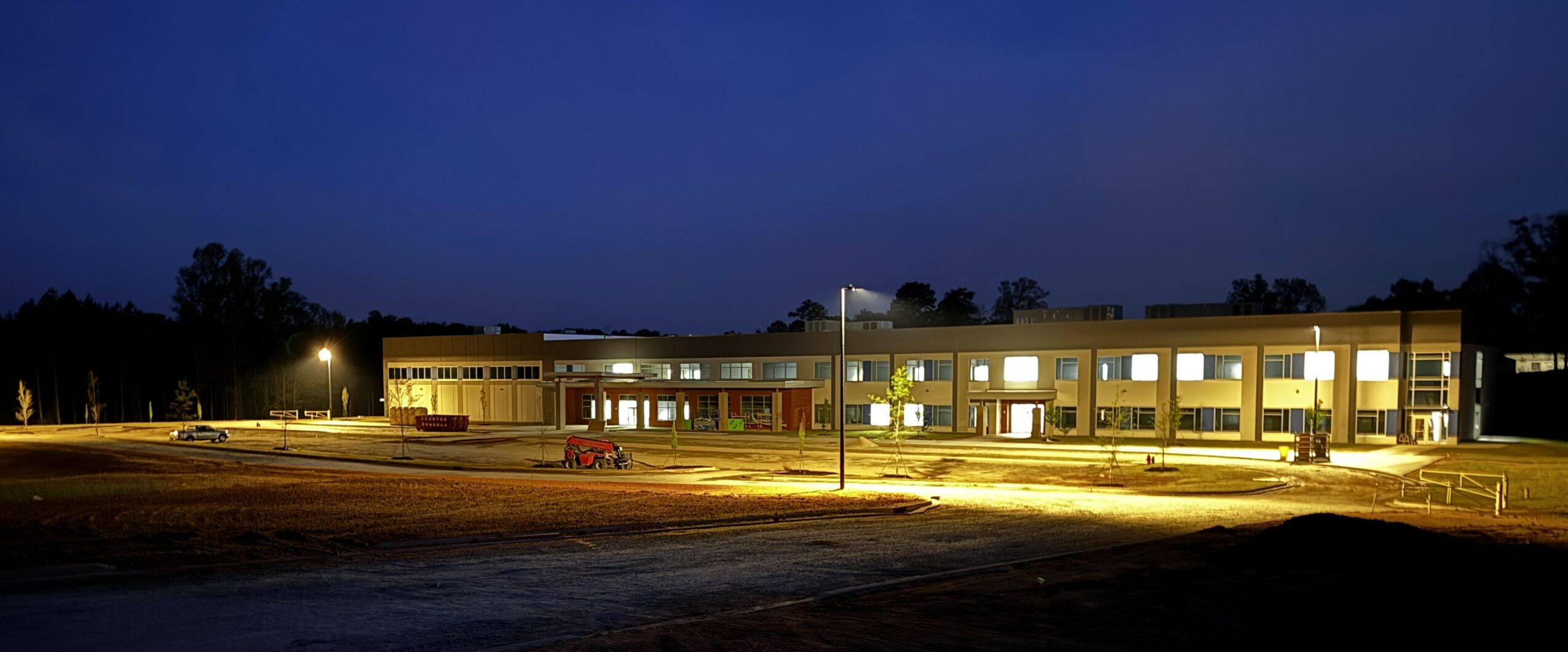 Building with illuminated windows at night, surrounded by an empty parking lot and trees in the background.