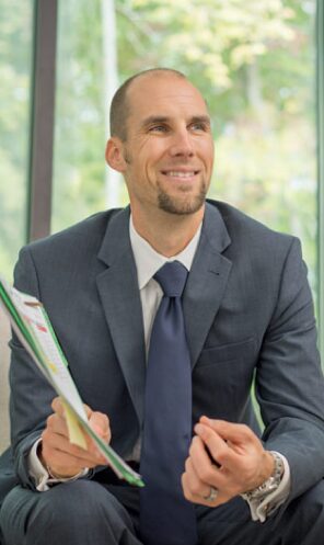Man in suit holding papers, smiling, sitting indoors with a window and green trees outside.