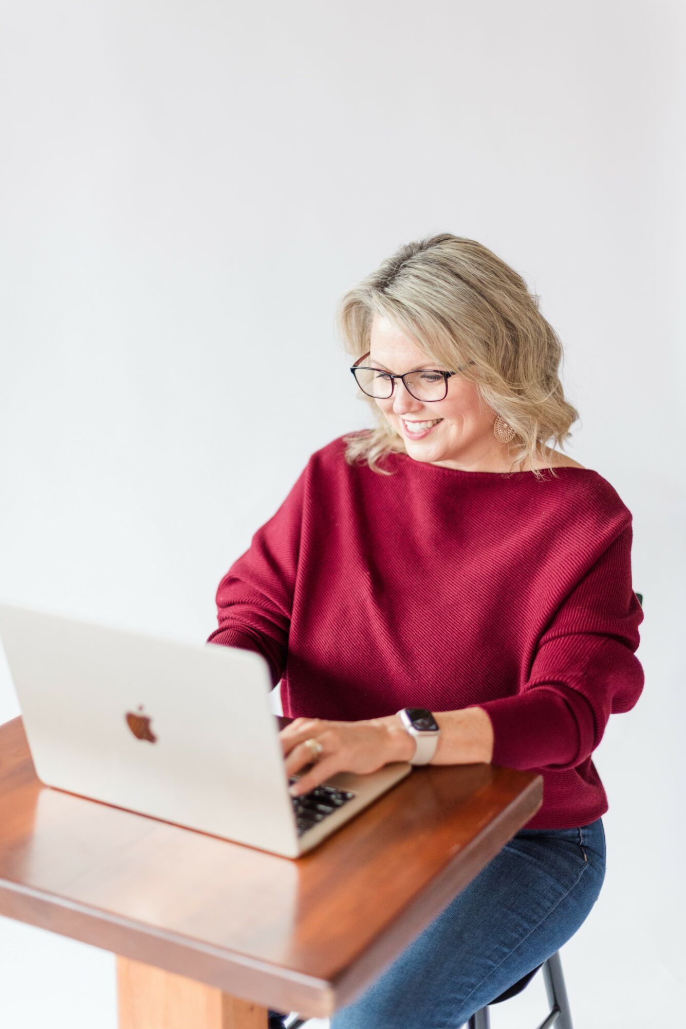 Woman with glasses and blonde hair working on a laptop at a wooden table.